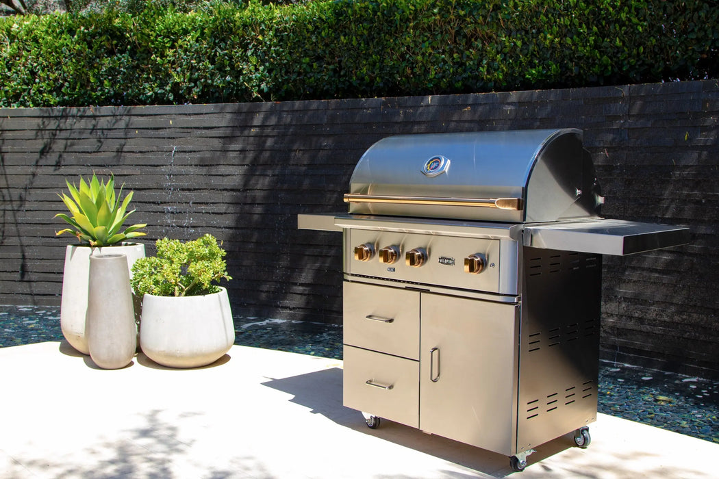 Wildfire Ranch Pro 36" gas grill in stainless steel with brushed bronze Designer Finish hardware on its cart on a patio against a dark wall, front view
