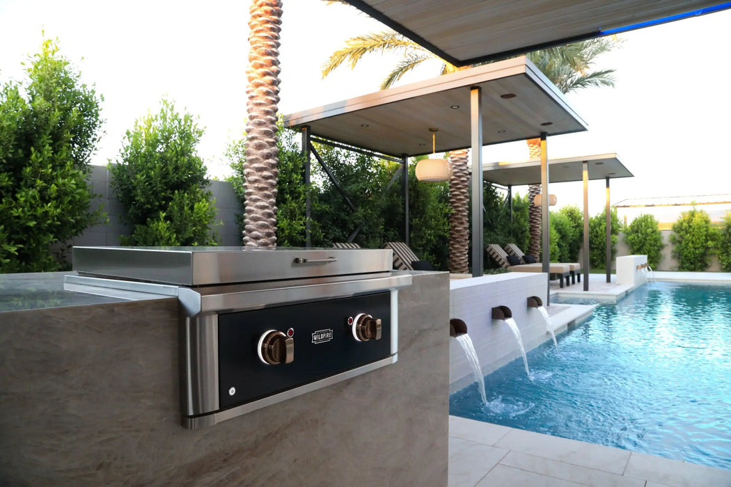 Wildfire Ranch Pro 30" gas griddle in black stainless steel with brushed bronze Designer Finish knobs, built into a stone island beside a pool, cover on
