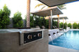 Wildfire Ranch Pro 30" gas griddle in black stainless steel with brushed bronze Designer Finish knobs, built into a stone island beside a pool, cover off